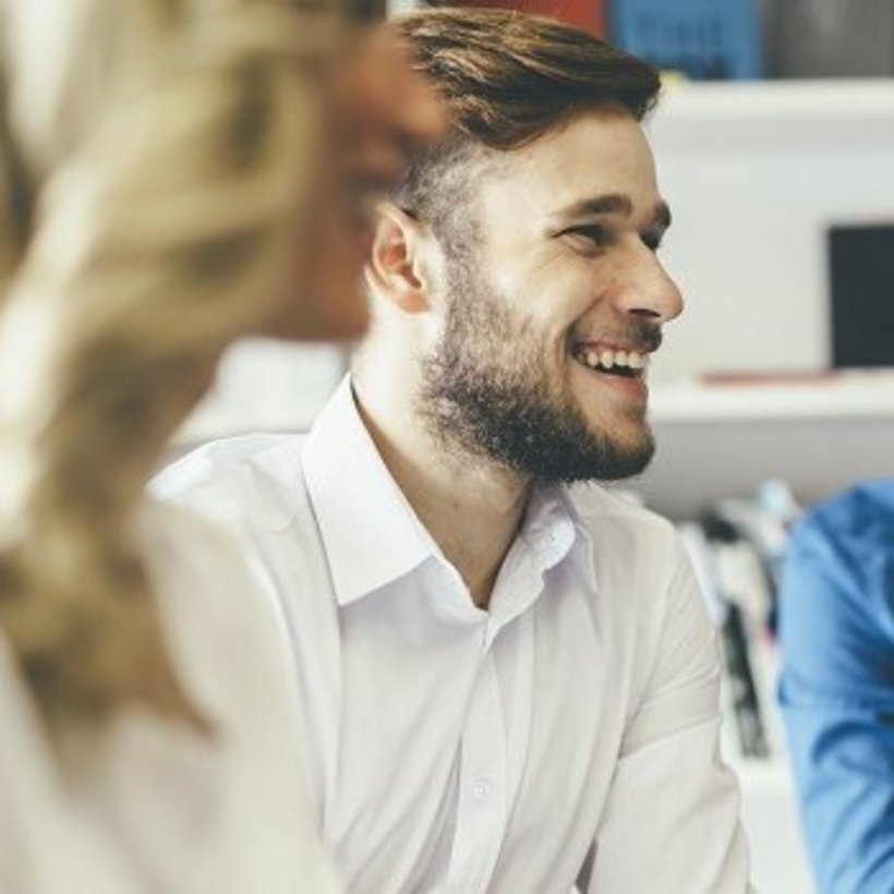 Drei junge Erwachsene sitzen in einem Büro oder Seminarraum. Ein Mann mit Bart und weißem Hemd lächelt, während ein Kollege daneben konzentriert auf einen Laptop schaut. Die Stimmung wirkt freundlich und produktiv.