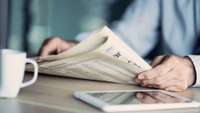 Person liest eine Zeitung an einem Tisch, neben der eine weiße Kaffeetasse und ein Tablet liegen. Das Bild kombiniert traditionelle Printmedien mit moderner Technik.