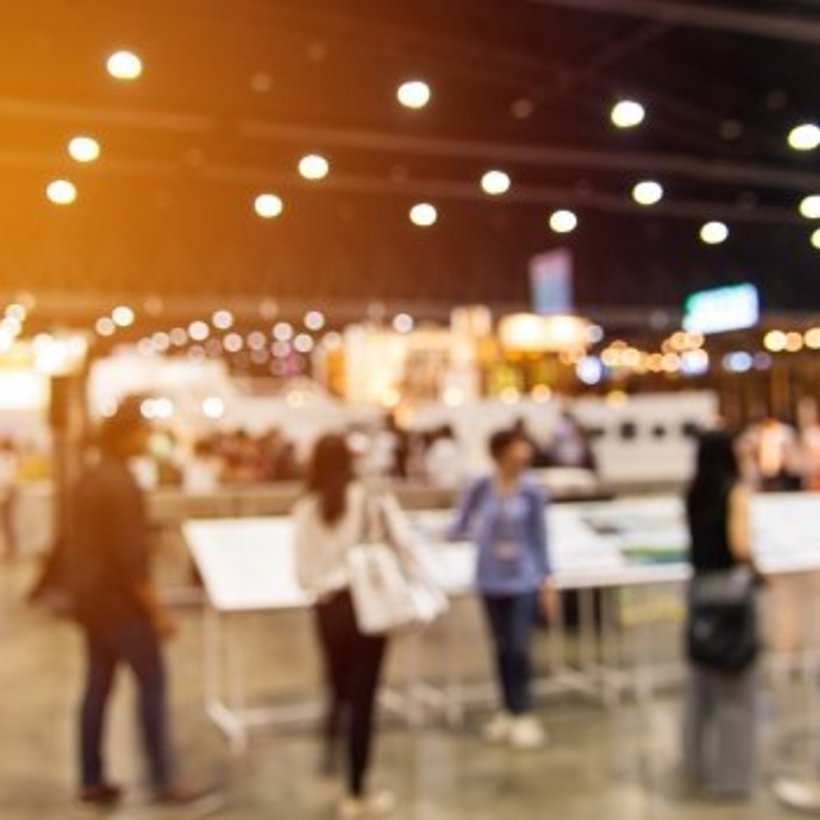 Unscharfe Aufnahme einer Messehalle mit vielen Menschen, die an Ständen und Infotafeln vorbeigehen. Helles Licht und warme Farben erzeugen eine lebendige, geschäftige Atmosphäre.