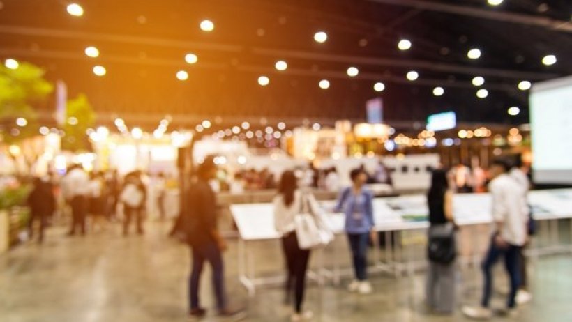 Unscharfe Aufnahme einer Messehalle mit vielen Menschen, die an Ständen und Infotafeln vorbeigehen. Helles Licht und warme Farben erzeugen eine lebendige, geschäftige Atmosphäre.