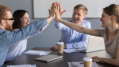 Vier Personen sitzen an einem Konferenztisch und geben sich lachend ein gemeinsames High-Five. Auf dem Tisch liegen Laptops, Notizbücher und Kaffeebecher, die Stimmung wirkt motiviert und positiv.