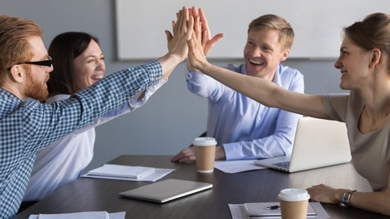 Vier Personen sitzen an einem Konferenztisch und geben sich lachend ein gemeinsames High-Five. Auf dem Tisch liegen Laptops, Notizbücher und Kaffeebecher, die Stimmung wirkt motiviert und positiv.