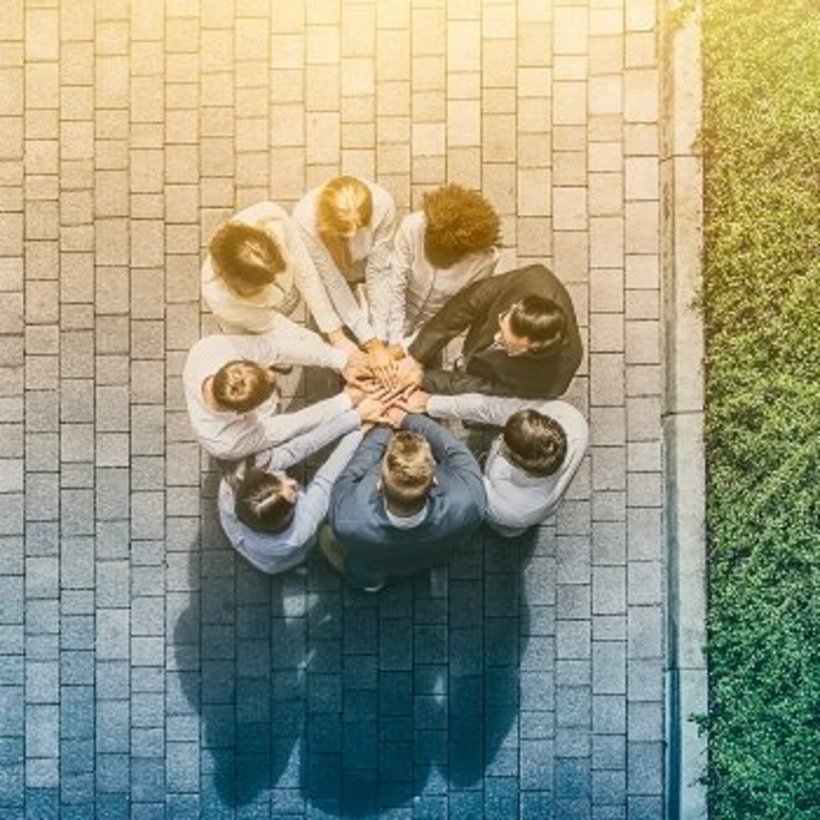 Luftaufnahme einer Gruppe von acht Personen, die im Kreis auf einem gepflasterten Platz stehen und ihre Hände in der Mitte übereinanderlegen. Rechts im Bild ist eine Grünfläche zu sehen, das Licht wirkt warm und freundlich.