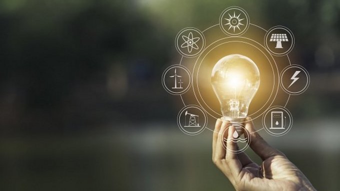 Hand hält eine leuchtende Glühbirne im Freien. Um die Birne sind grafisch Symbole für erneuerbare Energien und Nachhaltigkeit eingeblendet, darunter Sonne, Windkraft, Wasserkraft und Strom.