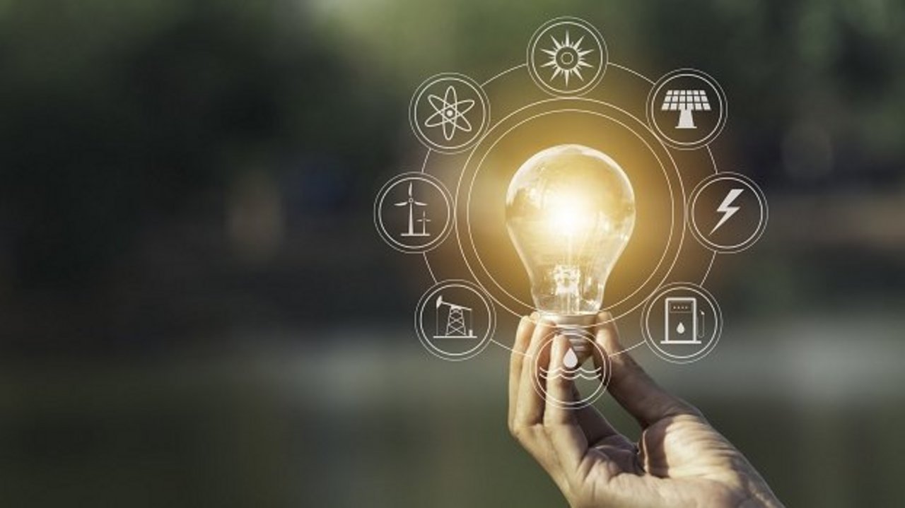 Hand hält eine leuchtende Glühbirne im Freien. Um die Birne sind grafisch Symbole für erneuerbare Energien und Nachhaltigkeit eingeblendet, darunter Sonne, Windkraft, Wasserkraft und Strom.
