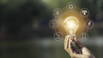 Hand hält eine leuchtende Glühbirne im Freien. Um die Birne sind grafisch Symbole für erneuerbare Energien und Nachhaltigkeit eingeblendet, darunter Sonne, Windkraft, Wasserkraft und Strom.