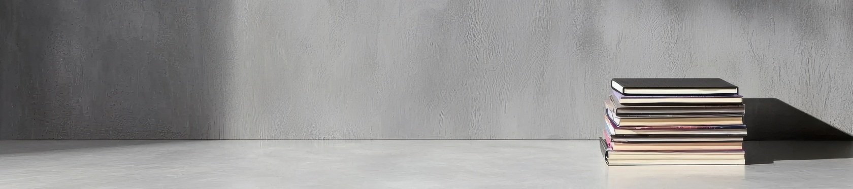 Ein Stapel Bücher liegt auf einem hellen Boden vor einer grauen Wand, auf die schräg Licht und Schatten fallen.
