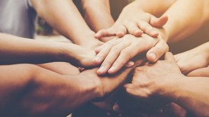 Mehrere Hände von unterschiedlichen Personen liegen übereinander in der Mitte, als Symbol für Zusammenhalt, Teamgeist und Kooperation.