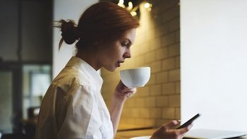 Junge Frau mit rotem Haar sitzt an einem Tisch, hält eine weiße Tasse in der Hand und schaut konzentriert auf ihr Smartphone. Im Hintergrund sind eine helle Wand und Fliesen zu sehen, die Szene wirkt wie in einem Café oder einer modernen Küche.