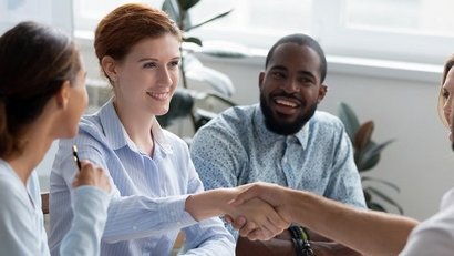 Vier Personen sitzen in einem hellen Büro an einem Tisch. Eine Frau mit hellem Hemd schüttelt lächelnd die Hand eines Gegenübers, während zwei weitere Teammitglieder daneben freundlich zusehen. Die Szene wirkt kooperativ und positiv.