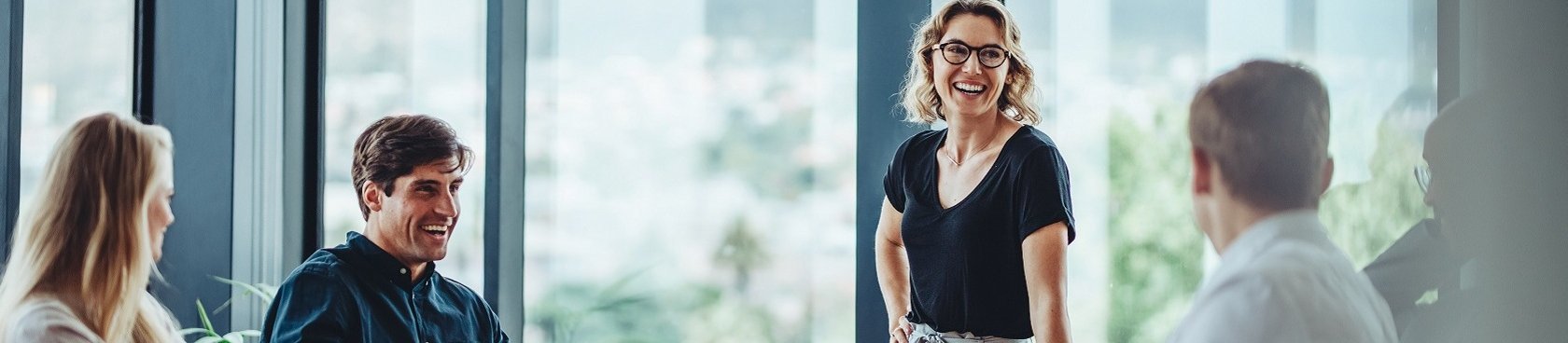 Ein Besprechungsraum gefüllt mit vier Menschen an einem Tisch. Eine Frau am Tischende steht und hat die Aufmerksamkeit der anderen auf sich gerichtet. 