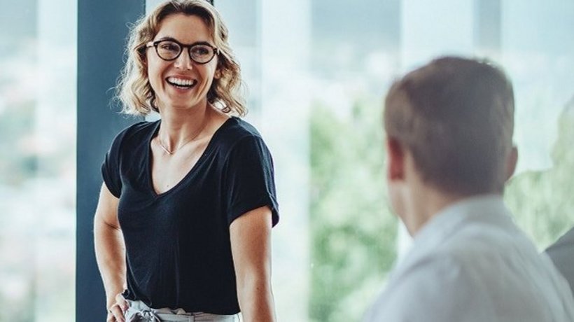 Ein Besprechungsraum gefüllt mit vier Menschen an einem Tisch. Eine Frau am Tischende steht und hat die Aufmerksamkeit der anderen auf sich gerichtet. 