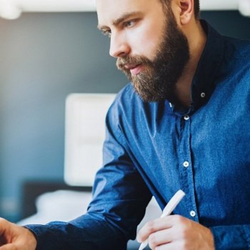 Mann mit Bart und blauem Hemd arbeitet konzentriert an einem Laptop, in der Hand hält er einen Stift. Neben ihm steht eine weiße Kaffeetasse, im Hintergrund fällt Tageslicht durch ein Fenster.
